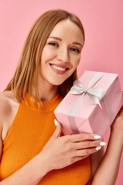 A woman in an orange top joyfully holds a pink gift box. — Stock Photo