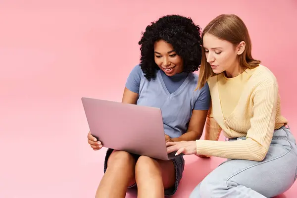 Two diverse women in casual attire are sitting on the ground, focused on a laptop. — Stock Photo