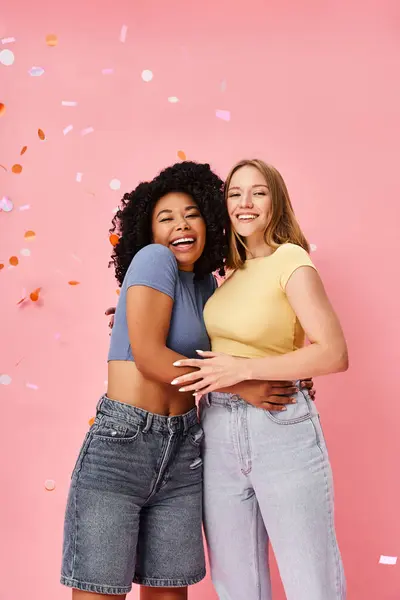 Two attractive diverse women in casual attire hugging each other in front of a pink background. — Stock Photo