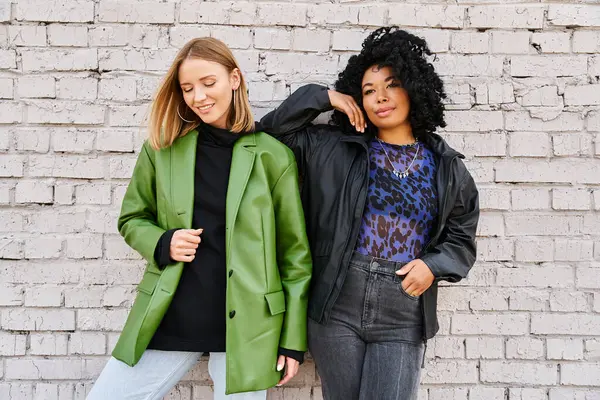 Two women of different ethnicities in casual clothing stand next to each other in front of a brick wall. — Stock Photo