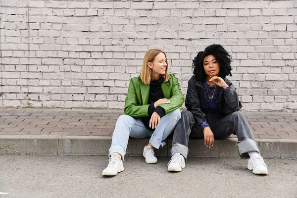 Deux femmes diverses en tenue décontractée assises sur un trottoir près d'un mur de briques. — Photo de stock
