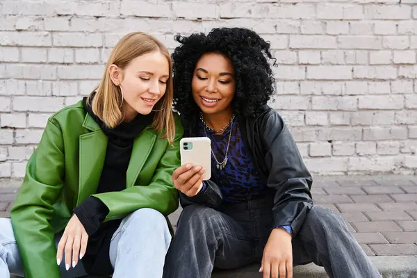 Two diverse women in casual attire sitting on ground, engrossed in cell phone. — Stock Photo