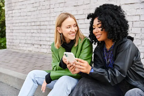 Two women in casual attire sitting on steps, engrossed in a cell phone. — Stock Photo