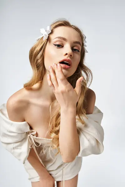 Young blonde woman exudes elegance, posing gracefully in a white dress with hands delicately framing her face in a studio. — Stock Photo