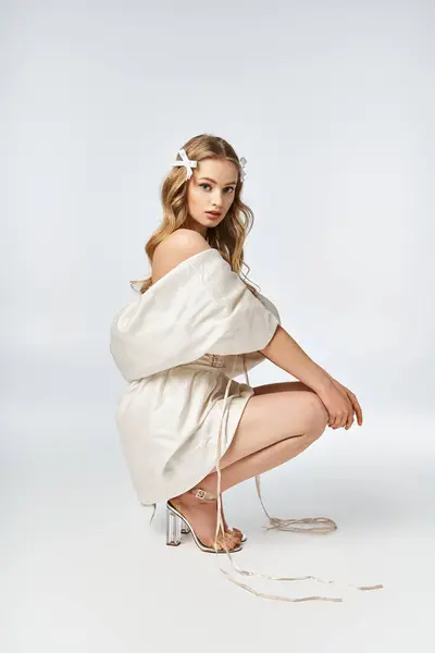 A young, blonde woman kneels gracefully in a flowing white dress in a studio setting. — Stock Photo