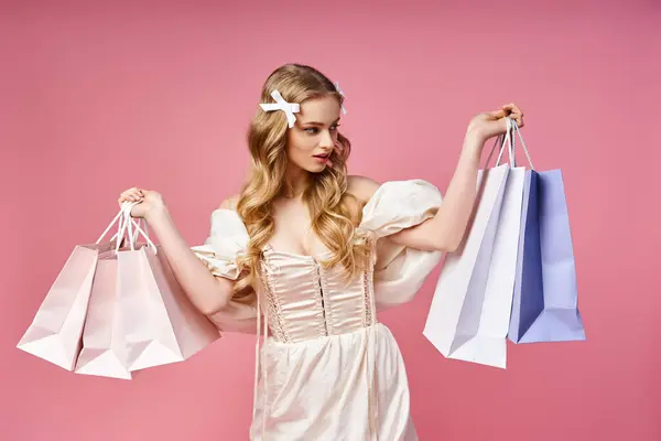 Una joven rubia con un vestido blanco sostiene bolsas de compras en un ambiente de estudio. - foto de stock