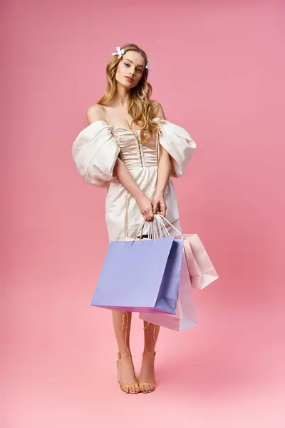 Una joven rubia con un vestido blanco impresionante sostiene múltiples bolsas de compras en un entorno de estudio. - foto de stock
