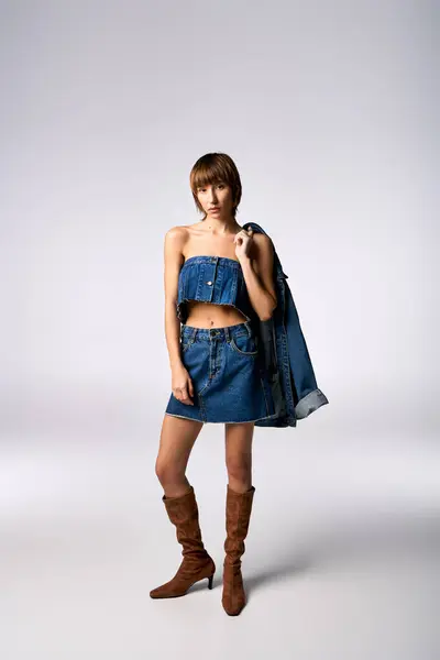 Une jeune femme confiante aux cheveux courts pose dans une robe en denim et des bottes pour un shooting de mode en studio. — Photo de stock