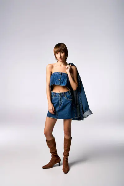 A confident young woman with short hair strikes a pose in a denim dress and boots for a fashion shoot in a studio. — Stock Photo