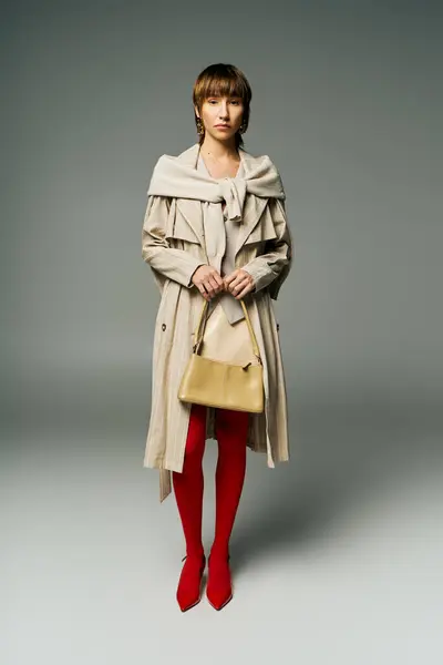 A stylish young woman with short hair poses in a trench coat and bright red tights in a studio setting. — Stock Photo