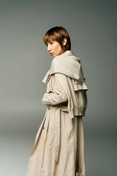 Une jeune femme aux cheveux courts se tient en confiance dans un trench-coat sur un fond gris uni. — Photo de stock