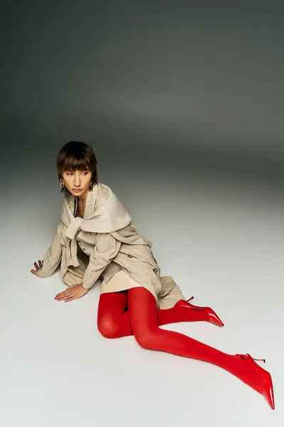 Une jeune femme aux cheveux courts dégage un charme séduisant dans un trench-coat rouge et des bas assortis dans un décor studio. — Photo de stock