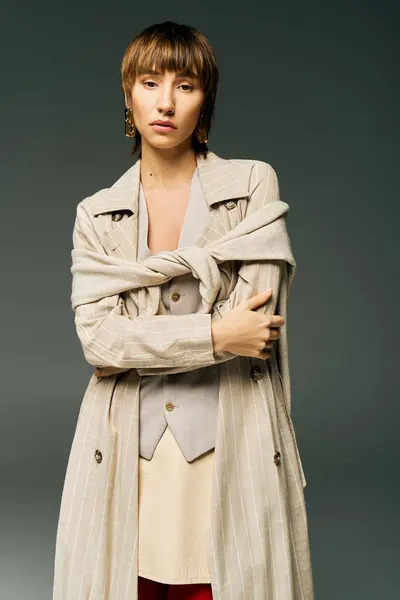 A young woman with short hair poses confidently in a trench coat in a studio setting, exuding elegance and poise. — Stock Photo