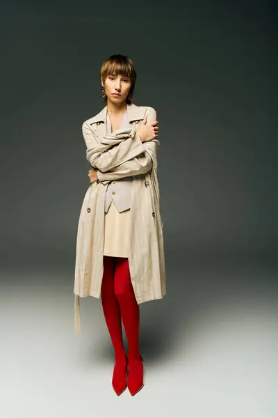 A young woman with short hair poses in a studio wearing a trench coat and vibrant red tights, exuding sophistication and style. — Stock Photo