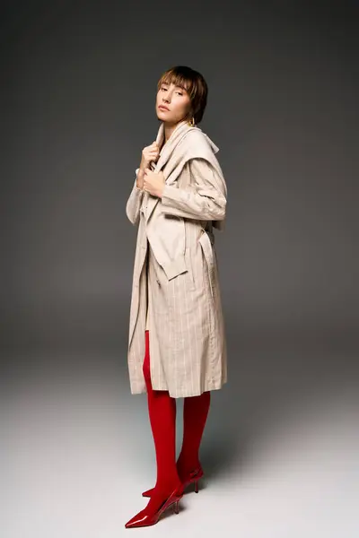 Молода жінка з коротким волоссям впевнено штовхається в стильне тренч пальто і яскраві червоні черевики в обстановці студії. — стокове фото