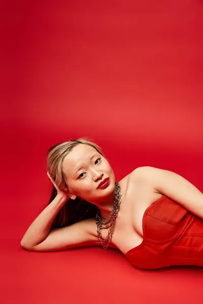 Una atractiva mujer asiática en un vestido rojo descansa graciosamente en el suelo. - foto de stock