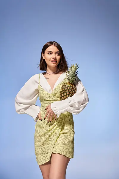 Beautiful young woman in a flowing dress gracefully holds a ripe pineapple in a vibrant studio setting. — Stock Photo