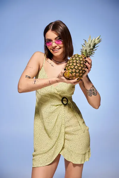 A brunette woman in a yellow dress joyfully holds a pineapple in a studio setting. — Stock Photo