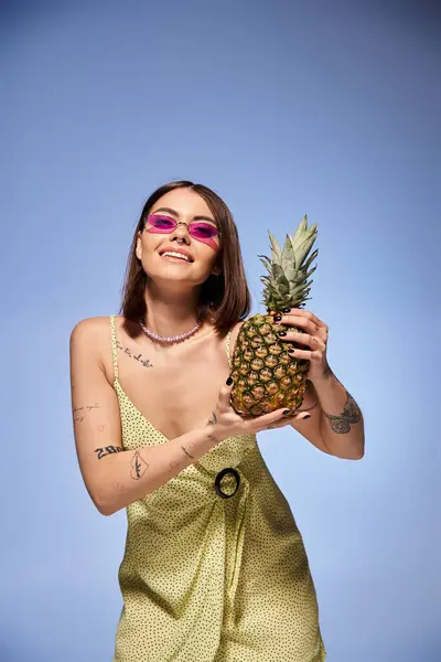 A brunette young woman elegantly holds a pineapple while wearing a vibrant yellow dress in a studio setting. — Stock Photo