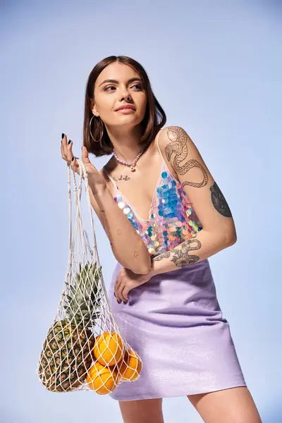 Una donna bruna con tatuaggi tiene una borsa piena di un assortimento di frutta fresca, in mostra un mix di natura e arte. — Foto stock