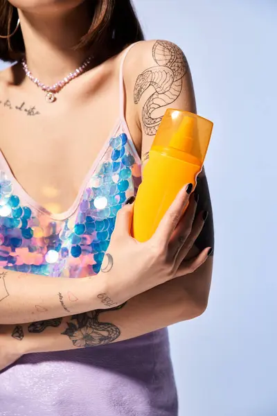 A young woman with tattoos, brunette hair, holding a yellow sunscreen in a sunlit studio setting. — Stock Photo