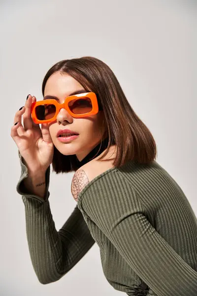 Una joven con el pelo moreno mostrando un suéter verde vibrante y elegantes gafas de sol de color naranja en un entorno de estudio. — Stock Photo