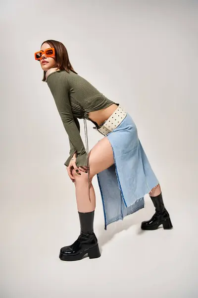 Une jeune femme élégante aux cheveux bruns pose en toute confiance dans une tenue chic comprenant une jupe et des bottes dans un cadre de studio. — Photo de stock