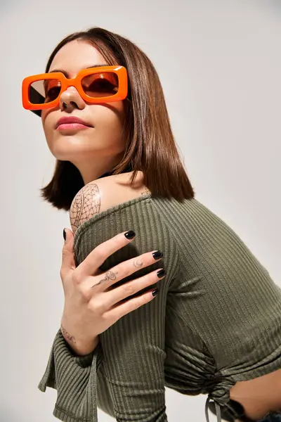 Una giovane donna chic con i capelli castani che indossa occhiali da sole rossi e un top verde in un ambiente da studio. — Foto stock