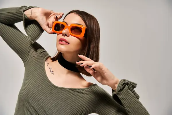 Uma jovem mulher com cabelo morena vestindo uma camisa verde e óculos de sol laranja posa em um estúdio. — Fotografia de Stock