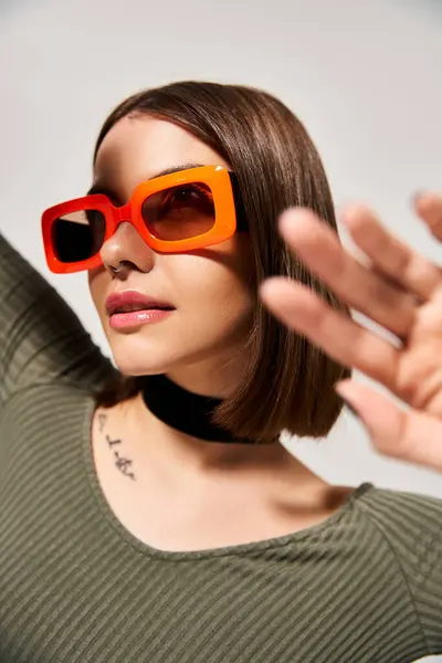 A stylish young woman with brunette hair wearing orange sunglasses and a green shirt in a studio setting. — Stock Photo
