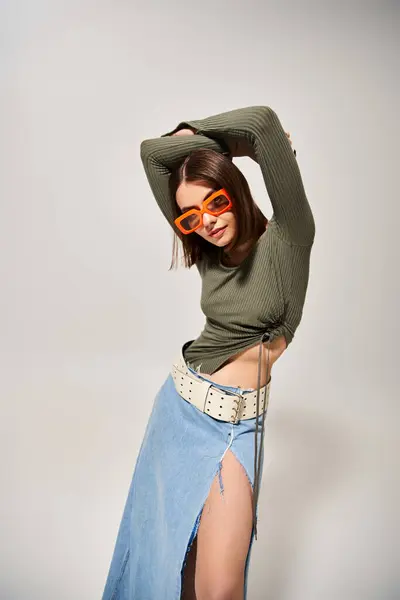 Una mujer joven y elegante con el pelo moreno posa con confianza en una falda y gafas de sol en un entorno de estudio profesional. - foto de stock