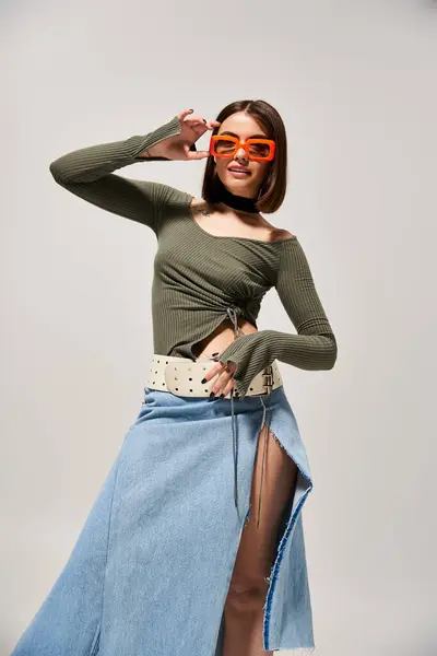 Una joven de moda con el pelo moreno posando con confianza en una falda y gafas de sol en un entorno de estudio. - foto de stock