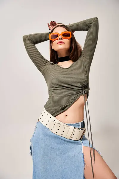 Une jeune femme élégante aux cheveux bruns pose en toute confiance dans une jupe et des lunettes de soleil pour un portrait en studio. — Photo de stock