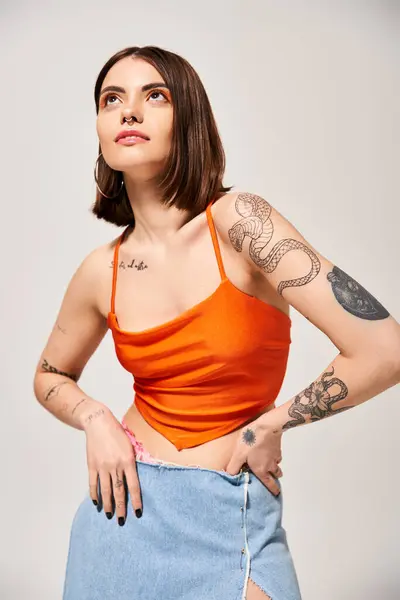 Una mujer joven con el pelo moreno golpeando una pose segura, las manos en las caderas, en una vibrante parte superior naranja en un entorno de estudio. - foto de stock