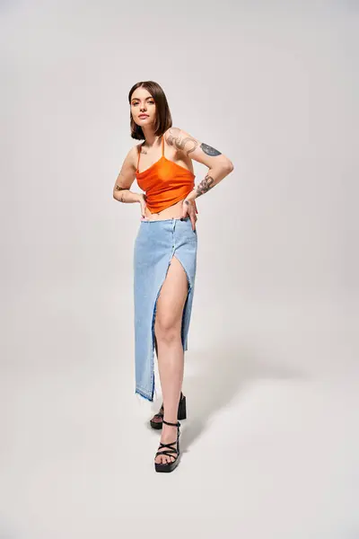 A stylish young woman with brunette hair stands gracefully in a studio wearing an orange top and a flowing blue skirt. — Stock Photo