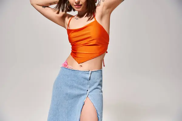 A young woman with brunette hair wearing an orange top and a blue skirt poses in a studio setting. — Stock Photo