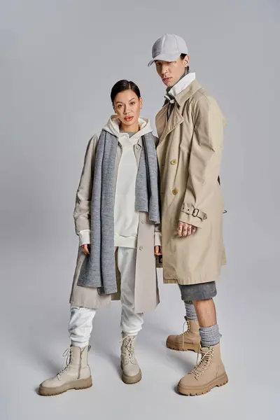 Um jovem casal elegante está lado a lado em casacos de trincheira na moda, exalando confiança e elegância contra um pano de fundo cinza. — Fotografia de Stock