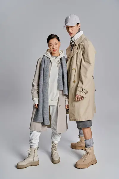 A stylish young couple stands side by side in trendy trench coats, exuding confidence and elegance against a grey backdrop. — Stock Photo