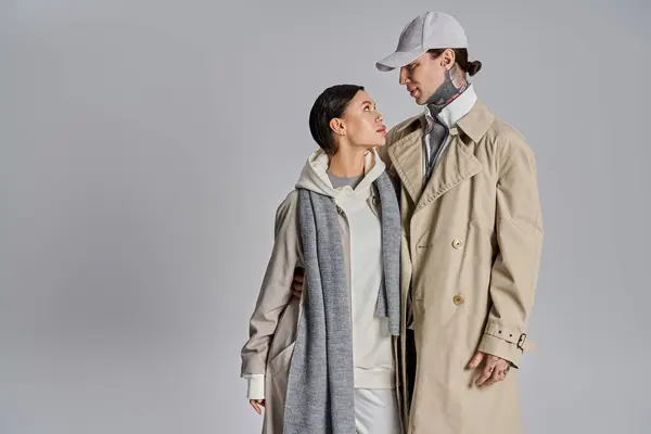 Un joven hombre y una mujer se paran uno al lado del otro, ambos llevando abrigos de trinchera, exudando estilo y confianza en un ambiente de estudio sobre un fondo gris.. - foto de stock