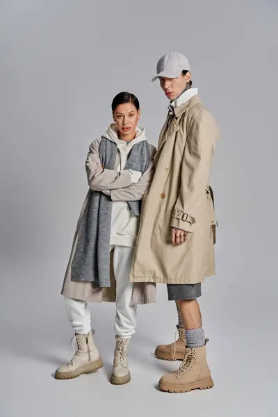 A young, stylish man and woman standing next to each other in trench coats against a grey studio backdrop. — Stock Photo