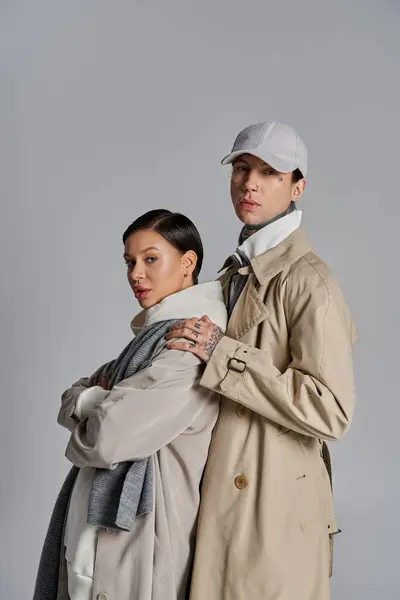 A young man and woman elegantly stand side by side in trench coats in a studio against a grey background. — Stock Photo