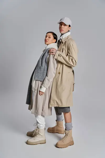 A young stylish couple stands side by side in trench coats, exuding sophistication and grace in a studio against a grey background. — Stock Photo