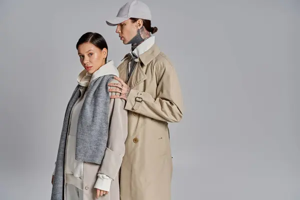 A young stylish couple standing together in trench coats in a studio against a grey background. — Stock Photo