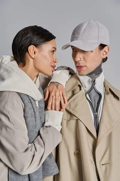 A young man and woman in trench coats stand together in a studio, their bodies angled towards each other on a grey background. — Stock Photo
