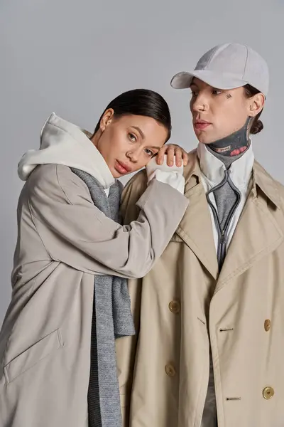 A young man and woman stand elegantly side by side in trench coats, showcasing their stylish attire in a studio setting against a grey background. — Stock Photo