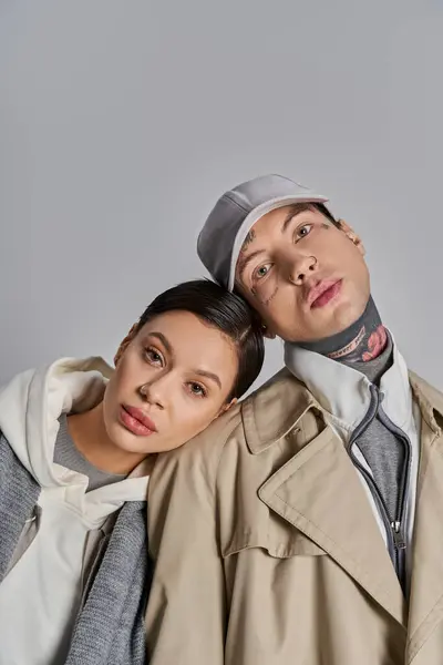 Un joven hombre y una mujer se paran con estilo uno al lado del otro en abrigos de trinchera, exudando un aire de sofisticación urbana sobre un fondo gris del estudio. - foto de stock