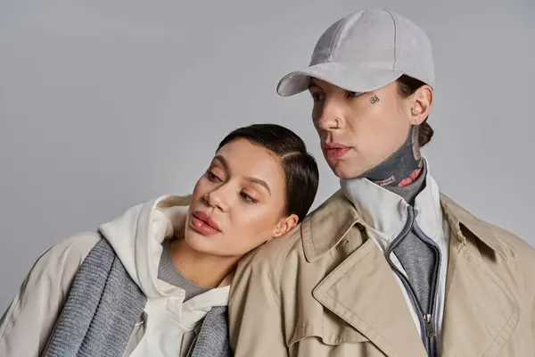 Ein junges stylisches Paar in Trenchcoats steht dicht an dicht in einem Atelier vor grauem Hintergrund.. — Stockfoto