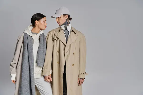 A young man and woman stand side by side in trench coats, exuding elegance and style against a grey studio backdrop. — Foto stock
