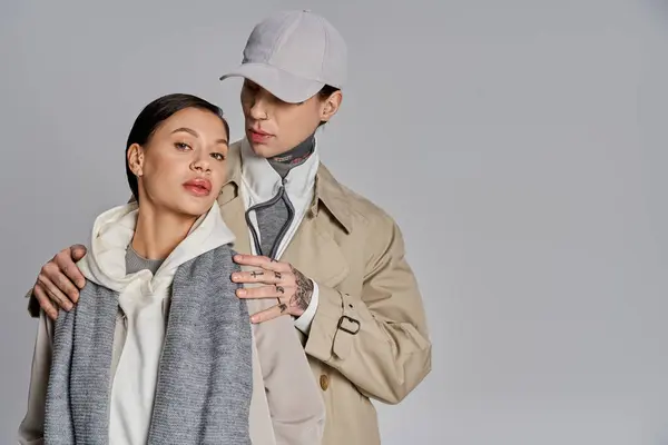 A young man and woman, both stylishly dressed in trench coats, stand side by side in a studio against a grey background. — Stock Photo