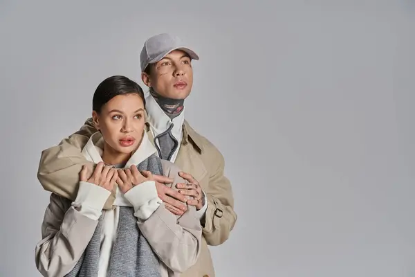 A stylish young man and woman wearing trench coats stand side by side in a studio against a grey background. — Stock Photo